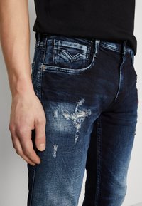 Mörkblå denimjeans med urblekta fläckar och slitna detaljer. Har fem fickor och kontrasterande sömmar. Hand placerad på låret.