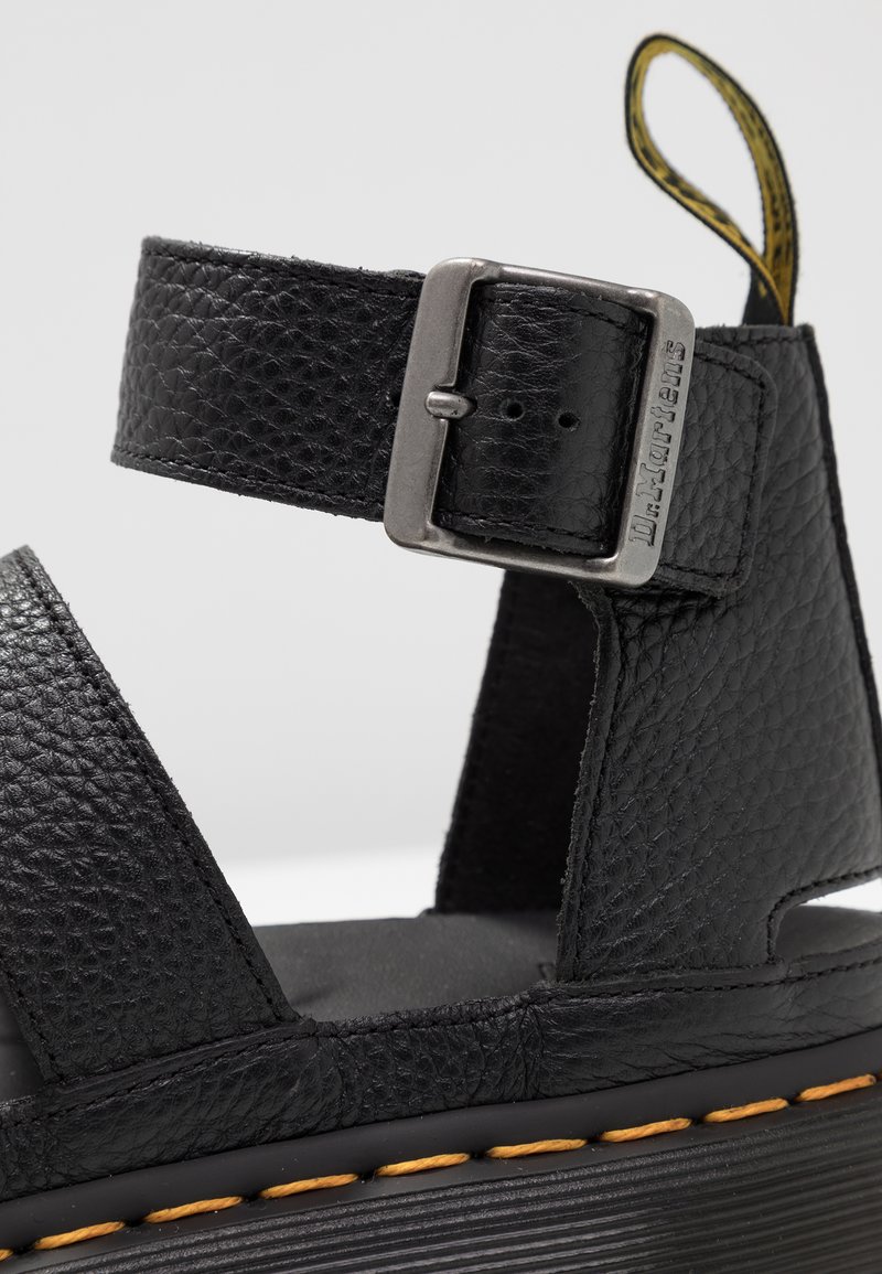 Black leather sandal featuring a textured strap, silver metal buckle, and yellow accent stitching on the sole. Rounded design with adjustable fit.