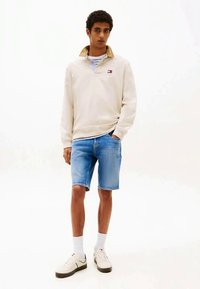 Νέος άντρας στέκεται φορώντας ένα μπεζ πουλόβερ με γιακά, ριγέ πουκάμισο από κάτω, μπλε τζιν σορτς, λευκές κάλτσες και λευκά αθλητικά παπούτσια.