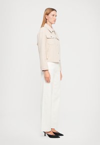 Giacca beige corta con due tasche sul petto e bottoni in argento, abbinata a pantaloni bianchi a gamba larga e décolleté neri a punta.