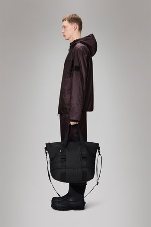 Sleek black duffel bag with padded texture, dual handles, adjustable shoulder strap; worn by a figure in a dark purple waterproof jacket and boots.