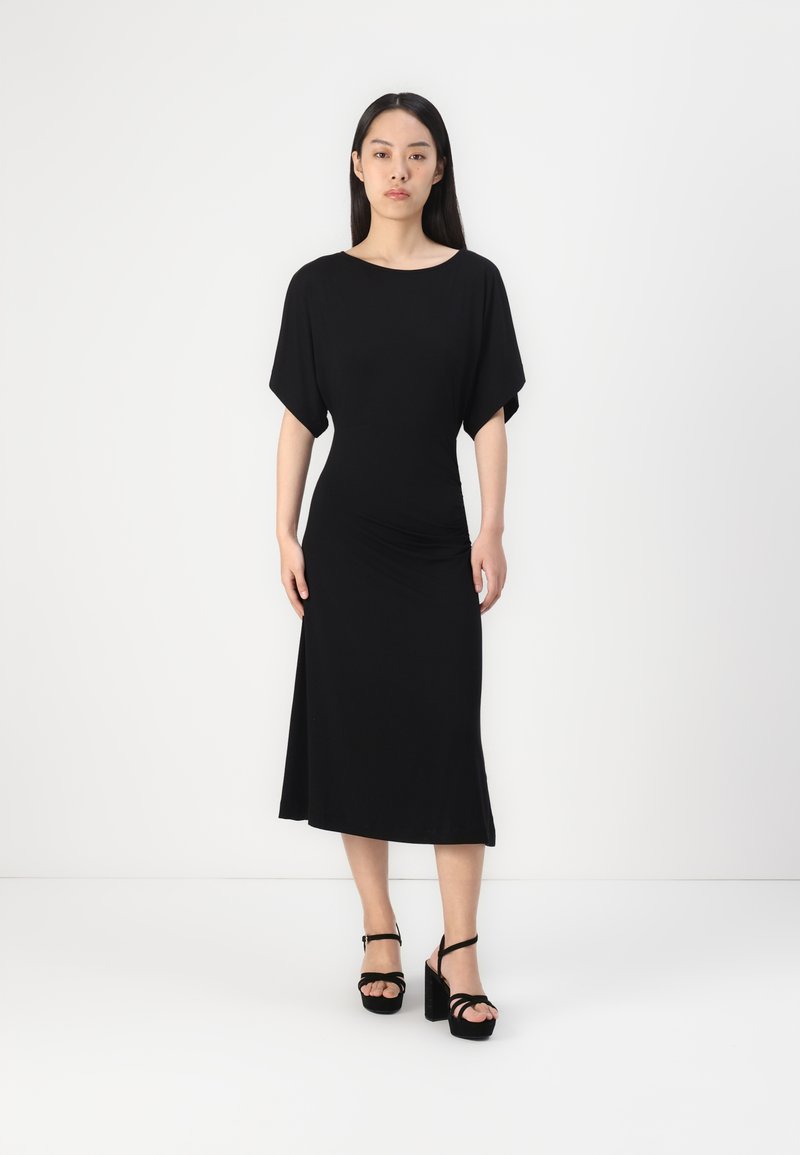Zwarte jurk tot de knie met korte vleermuismouwen, gemaakt van een gladde stof. Gecombineerd met zwarte platform sandalen met enkelbandjes.