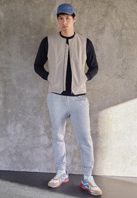 Beige zip-up vest over een zwart langemouwen shirt, gecombineerd met grijze joggingbroeken en meerkleurige sneakers. Een blauwe pet maakt de outfit af.