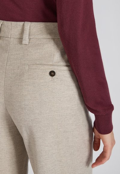 Lichtbeige visgraatbroek met een gestructureerde afwerking, voorzien van een achterzak met een donkere knop. Bordeaurode lange mouwen top zichtbaar.