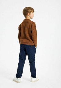 Brązowy sweter w kratę z okrągłym dekoltem, zestawiony z granatowymi spodniami i białymi trampkami. Sweter ma prążkowane mankiety i dół.