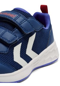 Niebieski but sportowy wykonany z siateczkowego materiału, z białym zygzakowatym wzorem, paskami na rzepy oraz teksturowaną podeszwą z fioletowymi akcentami i czerwonym uchwytem do ściągania.