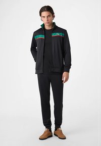 Sort træningsdragt med en lynlåstrøje med grønne og røde striber og logo, kombineret med matchende bukser. Inkluderer brune sneakers.