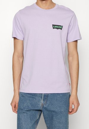 Mand iført lys lilla Levi's t-shirt med lille sort og grønt logo på brystet, kombineret med blå denimjeans.