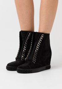 Czarne zamszowe botki na koturnie do kostki z zamkiem z przodu i metalowymi łańcuchami wzdłuż boków. Teksturowana czarna gumowa podeszwa z plecionymi detalami.