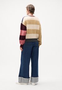 Strikkegenser med varierte striper i farger: krem, gull, burgunder og rosa, med et teksturert kabelmønster. Kombinert med vide denimbukser.