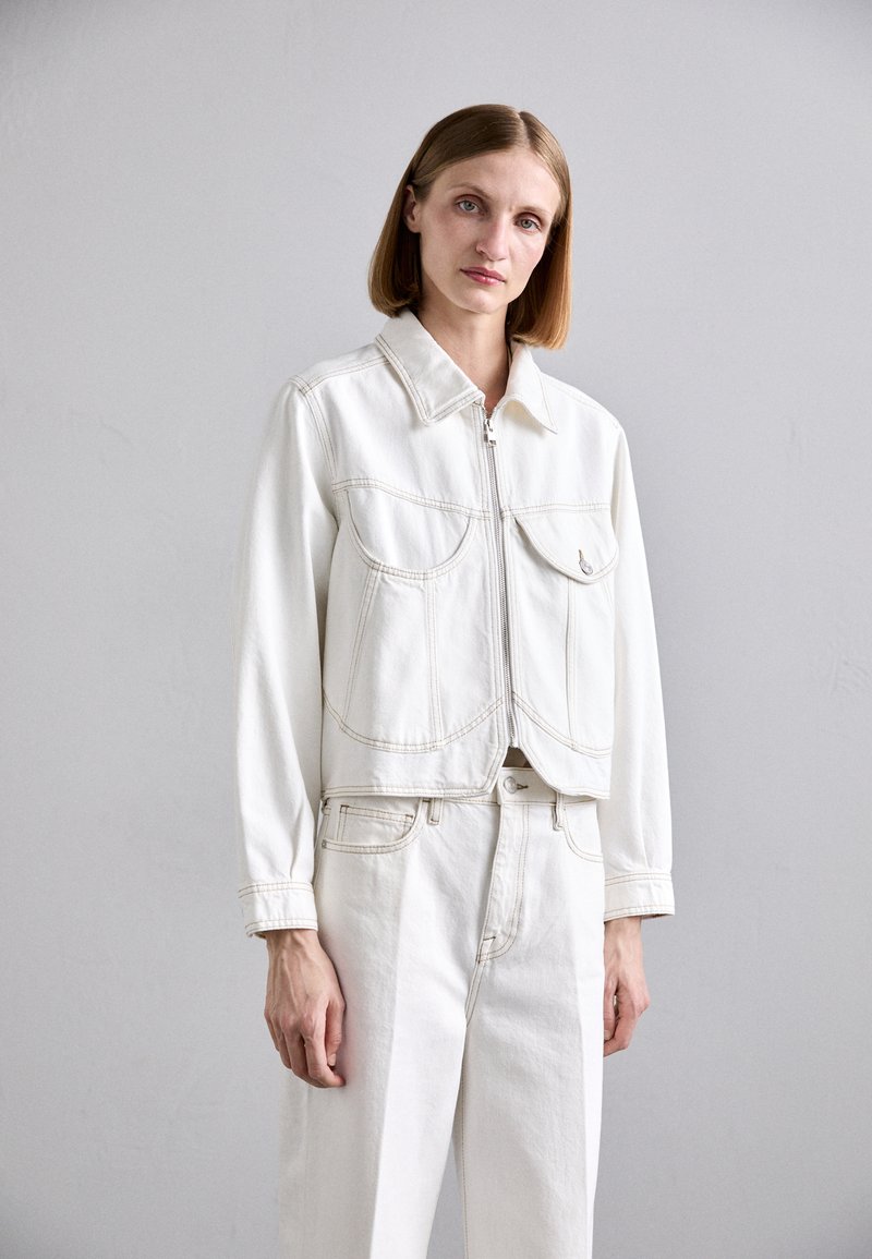 Weiße Jeansjacke mit zwei vorderen Taschen, versehen mit kontrastierenden Nähten und einem gekürzten Design. Modell trägt passende weiße Hosen.
