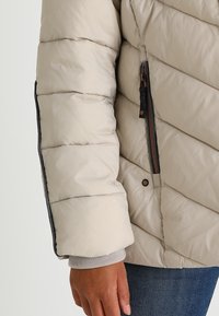 Jasnokremowa kurtka puchowa z ukośnym przeszyciem na rękawie, zamkiem błyskawicznym w kieszeni, szarym wykończeniem mankietu oraz ciemnymi akcentami wzdłuż szwów.