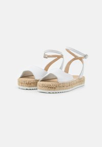 Bílé kožené espadrilky s řemínky kolem kotníků, pletené jutové platformové podešve a bílé gumové podrážky na bílém pozadí.