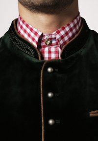 Dunkelgrünes Samtjackett mit braunen Besätzen, metallischen Knöpfen und komplizierter schwarzer Stickerei. Über einem rot-weiß karierten Hemd getragen.