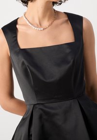 Abito di raso nero con scollatura quadrata, spalline larghe e corpetto aderente. La gonna plissettata aggiunge volume. Collana di perle e catena come accessorio.