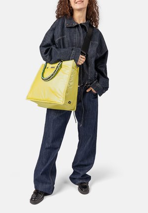 Vrouw draagt een donker denim jack en broek, houdt een grote gele draagtas met kleurrijke gevlochten handgreep vast, staand tegen een witte achtergrond.