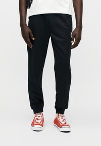 Μαύρο αθλητικό παντελόνι με εφαρμοστή σχεδίαση, ελαστική μέση και μανσέτες στους αστραγάλους. Συνδυασμένο με κόκκινα πάνινα παπούτσια με λευκές σόλες.