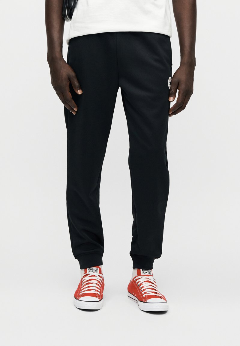 Sorte atletikbukser med tætsiddende design, elastisk talje og ankellukkede kanter. Kombineret med røde lærredssneakers med hvide såler.