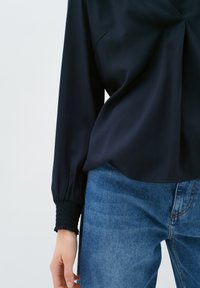 Blusa blu navy realizzata in tessuto liscio, con un design anteriore attorcigliato e polsini elasticizzati, abbinata a jeans in denim azzurro chiaro.