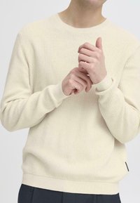 Maglione lavorato a maglia di colore beige chiaro con texture a costine, colletto a girocollo e polsini aderenti. Presenta un sottile dettaglio delle cuciture e una piccola etichetta del marchio sul lato.