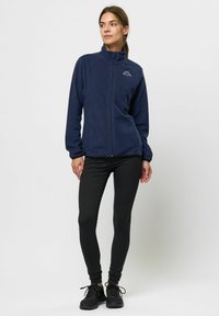 Marinfärgad fleecejacka med hög krage, heldragkedja och logotyp på bröstet, matchad med svarta leggings och svarta träningsskor. Slät textur.