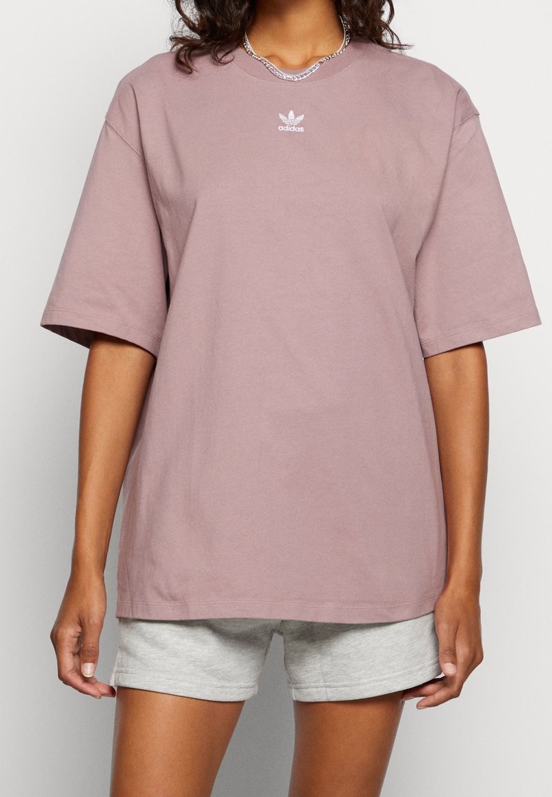 Hellrosa übergroßes Baumwoll-T-Shirt mit kurzen Ärmeln; weist ein kleines weißes Adidas-Logo auf der Brust und eine glatte Textur auf.