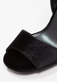 Chaussure noire en velours à bout ouvert présentant une large bride sur le dessus et une semelle lisse et brillante. Le design est minimaliste avec une texture douce.