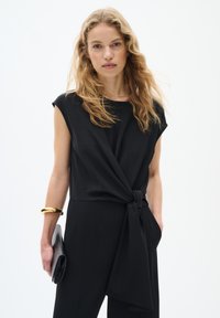 Combinaison noire avec un détail de lien noué à la taille, des manches larges et un tissu lisse et fluide. Les accessoires comprennent un bracelet en or et une pochette noire.