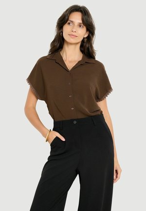 Vrouw draagt een bruin kort mouwen overhemd met knopen en kanten afwerking en zwarte hoog getailleerde broek, poseert met één hand in de zak.