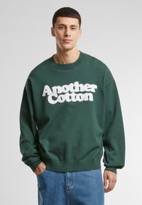 Sudadera de algodón verde con cuello redondo y ajuste oversized. Presenta texto blanco "Another Cotton" con una textura esponjosa en la parte delantera.
