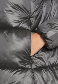 Černá péřová bunda s lesklým, prošívaným povrchem. Má kapsu na zip a manžetu na rukávu, které ukazují měkký, texturovaný materiál.