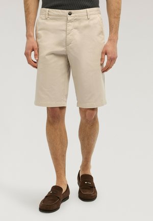 Mann trägt beige knielange Shorts und dunkelbraune Loafer, steht vor einem hellgrauen Hintergrund.