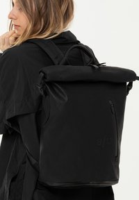 Zaino a rotolo nero realizzato in tessuto resistente, con tasca laterale con zip e dettagli testurizzati. Finitura liscia e spallacci regolabili.