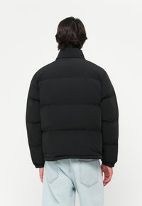 Doudoune noire avec un col montant, présentant un design matelassé et une coupe décontractée. Portée sur un jean bleu clair, montrant une texture lisse.