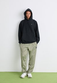 Mand iført sort Nike hættetrøje med hætten oppe, beige Nike træningsbukser og hvide sneakers, der står på et grønt gulv foran en hvid væg.