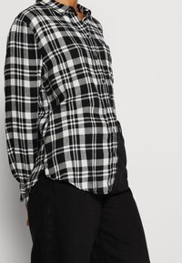 Camicia a quadri bianca e nera con maniche lunghe, orlo arrotondato e taschino sul petto. Realizzata in un tessuto testurizzato. Abbinata a pantaloni neri.