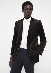 Brauner Cordblazer mit Revers, Einknopfverschluss und einer Brusttasche. Über einem weißen Hemd getragen. Dunkle Hosen.