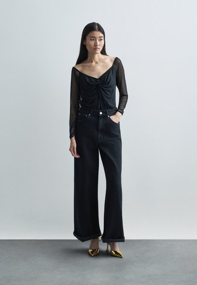 Top en maille noire à manches longues avec détail froncé, associé à un jean noir à taille haute et jambes larges, et des escarpins dorés à bout pointu.