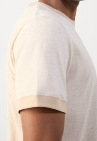 Beige kortærmet t-shirt med rund halsudskæring og kontrastfarvet lysebrun kant på ærmer og krave, lavet af blødt, tekstureret stof.