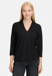 Pullover nera con scollatura a V e texture a coste, con maniche a tre quarti, indossato sopra jeans scuri. Design semplice e minimalista.