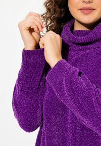 Lila plyschtröja med hög krage, ribbade muddar och textil struktur. Ärmarna är något oversized. Händer som greppar tröjan.