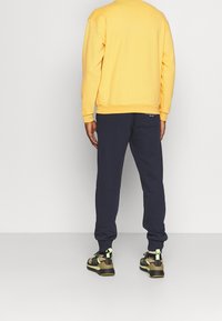 Felpa gialla a maniche lunghe, abbinata a pantaloni della tuta blu scuro; le scarpe presentano un motivo mimetico con accenti di verde neon.