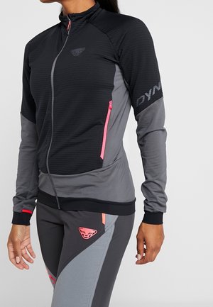 Frau trägt eine schwarz-graue Sportjacke mit pinken Reißverschlusstaschen und passende graue Leggings mit pinken Logopatches.