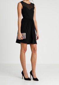 Robe noire sans manches avec un décolleté transparent, une taille froncée et une jupe évasée. Assortie d'une pochette à motifs et de talons hauts noirs.