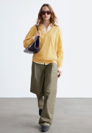 Kvinde iført gul sweater over hvid skjorte, olivengrønne bukser med vide ben, sorte sko, brune solbriller og bærende en sort skuldertaske, går fremad.