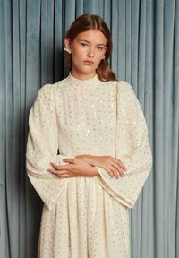 Mladá žena s dlhými vlasmi, ktorá má na sebe perlové náušnice a krémové šaty so vzorom z flitrov a nariasenými rukávmi, stojaca pred modrým závesom.