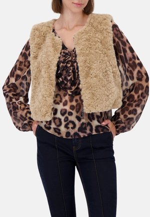 Beige Weste aus Kunstfell, verkürztes Design. Getragen über einer Bluse mit Leopardenmuster und Rüschenausschnitt. Dunkle Jeans runden das Outfit ab.