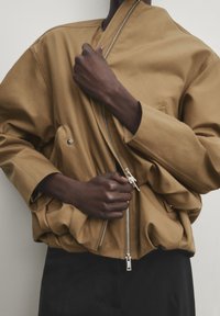 Veste marron surdimensionnée en tissu lisse, avec une fermeture éclair diagonale et une texture froncée à l'avant, associée à un pantalon foncé.