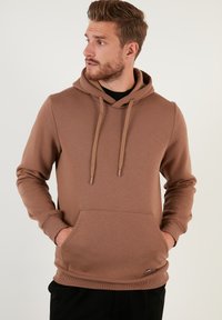 Sudadera marrón hecha de tela suave, con un bolsillo tipo canguro en la parte frontal, cordones ajustables y puños acanalados. Textura suave, corte casual.