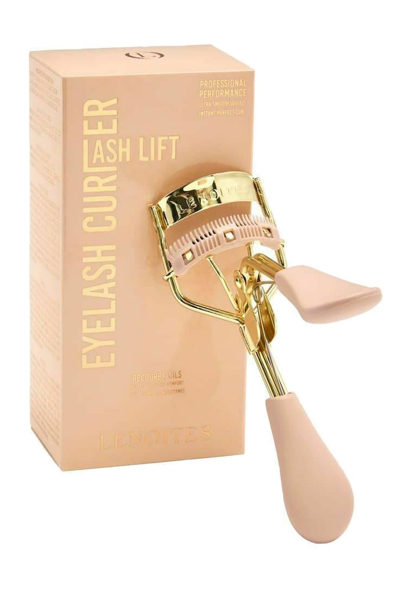 Recourbe-cils avec des accessoires en métal doré et des poignées en silicone rose. Accompagné d'une boîte rose clair portant un texte doré et des détails sur le produit.
